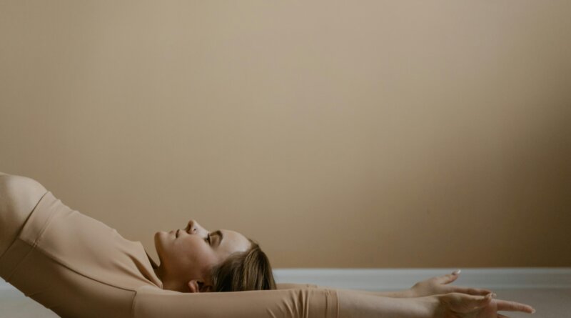 A woman lying flat on the floor in a relaxed position with arms extended overhead, wearing a beige outfit against a neutral background.